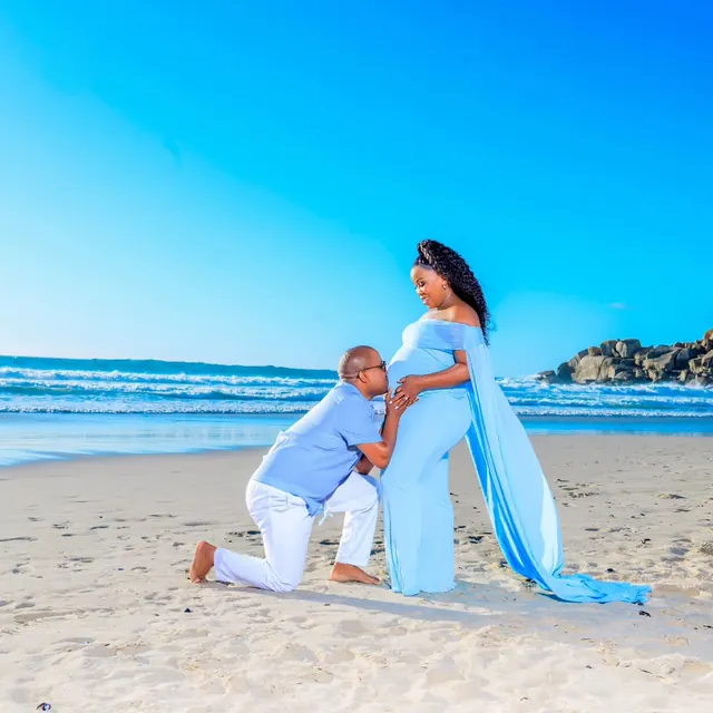 Maternity session, partner kneeling beside the bump, pale blue gown, beach